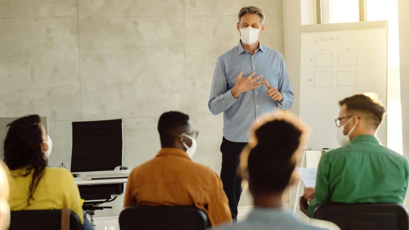 University professor wearing protective face mask while holding a class to group of students during coronavirus pandemic.