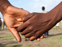 A closeup of two people holding hands, with a field in the background.