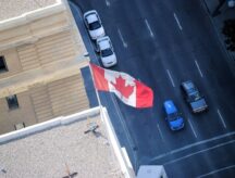 Looking down at a Canadian Maple Leaf flag waving in the wind on the top of a building and a few cars on the road below.