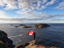 Woman standing on oceanside cliff holding Canadian flag.