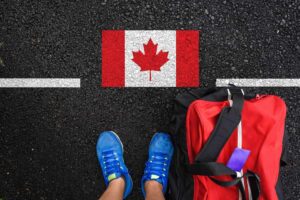 A person with a packed bag stands on a road with the Canadian flag in front of them.