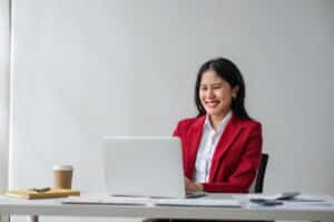 A woman in a red blazer smiles while sitting in front of a laptop.