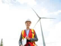 A enginerr with a hard hat smiling at the camera, with a windmill in the background of the image.