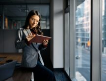 A woman in a suit starts at a tablet while standing next to a window