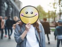 A person holds a cutout of a smiley face in front of their face.