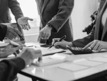 A group of people speaking around a conference room in black and white