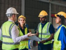 Factory workers wearing high visibility jackets and helmets talk to each other