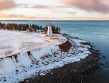 A red and white lighthouse standing along a coastline in the winter with a handful of homes behind it.