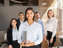 A woman in professional attire stands in front of other professionals, smiling.