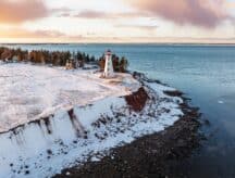 A red and white lighthouse standing along a coastline in the winter with a handful of homes behind it.