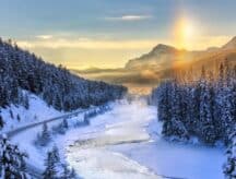 A beautiful winter landscape featuring mountains in the far distance, snowy trees on the left and right, and a frozen river in the centre. The sun is washing over the mountain tops.