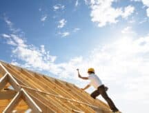 A construction worker in a yellow hard hat working on a finishing a roof. He is wielding a hammer.